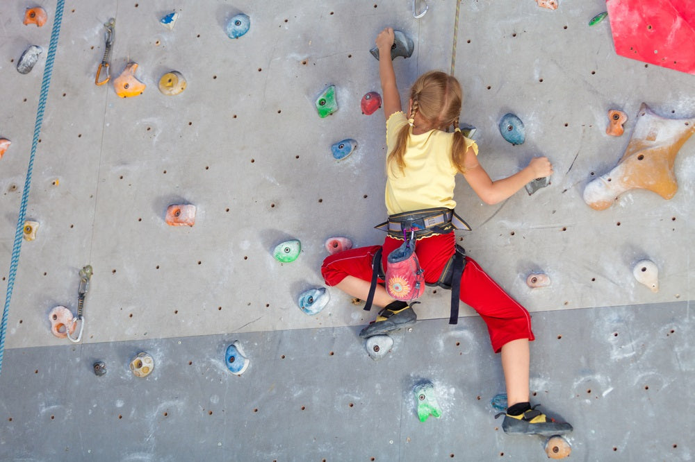 A young girl climbing an indoor rock wall wearing a safety harness, showcasing physical strength, coordination, and perseverance as part of child development activities.