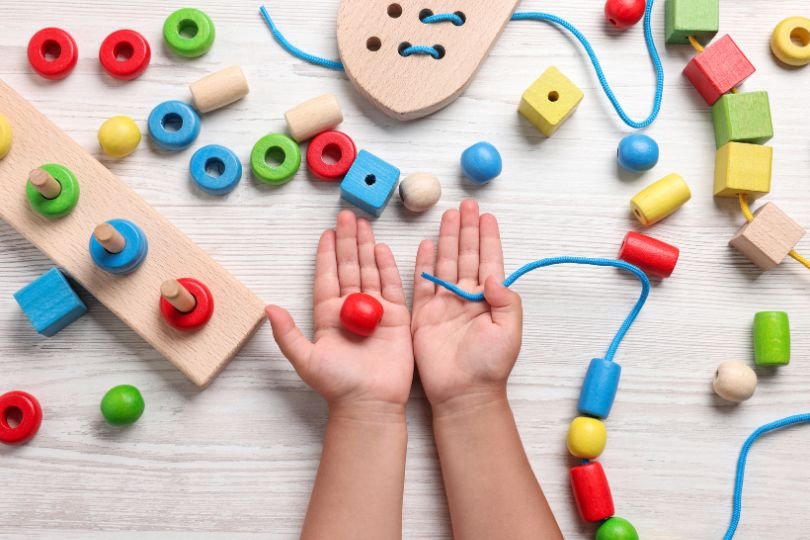 Hands of a child playing with colorful wooden Montessori threading beads and shapes, promoting fine motor skills and independent play.