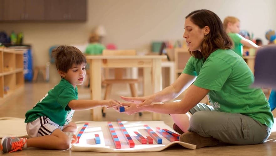 A toddler and teacher engaging with Montessori-style wooden blocks in a classroom, focusing on hands-on learning and fostering fine motor skills and cognitive development.